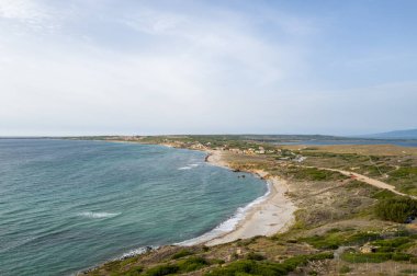 Capo San Marco, Sardinya 'nın turkuaz suları boyunca uzanan kumlu bir sahil manzarası. Temiz ve geniş bir gökyüzü altında küçük bir köy ve kıyı bitkisi..