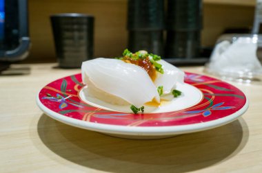 Turuncu balık yumurtası ve doğranmış yeşil soğanla kaplı iki parça şeffaf mürekkep balığı nigiri Japon restoranının soluk ahşap tezgahının üzerinde canlı kırmızı desenli bir tabakta servis edilir. Yumuşak iç aydınlatma parlak dokuyu vurgular ve...