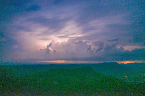 Karanlık çöktüğünde gölgeli tepelerin üzerinde büyük gök gürültülü bulutlar parıldar ve dramatik bir katman oluşturur. Sahnede ince siluetler, yumuşak ışık eğimleri ve manzara boyunca karamsar bir alacakaranlık atmosferi yer alıyor..