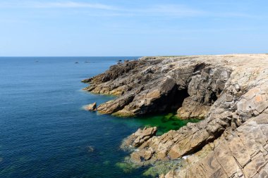 Engebeli kaya kayalıkları, Saint Pierre Quiberon 'daki Pointe Beg-An-Aud' daki sakin mavi okyanusun yanında, parlak, bulutsuz bir gökyüzünün altında canlı bir gelgit havuzuna iniyor..