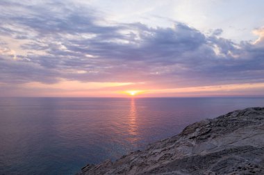 Gün batımı ışığı Stintino yakınlarındaki sakin denizi ve engebeli kayalıkları yumuşacık bulutlarla kaplar ve gökyüzünü lavanta ve altının gölgeleriyle boyar. Sakin atmosfer, Sardunya kıyı şeridinin doğal güzelliğini vurguluyor..