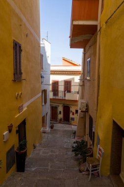 Sari ve bej cephelerle donanmis dik sokaklar, Castelsardo, Sardunya 'nin tarihi merkezinde, berrak bir gökyüzünün altinda, rustik panjurlar ve saksı bitkileri..