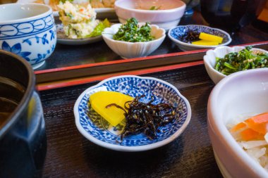 Close-up of yellow pickled radish and seasoned seaweed served in a blue and white ceramic dish, surrounded by assorted vegetable side dishes on a lacquered tray. Bright natural light enhances the fresh textures and vibrant presentation.