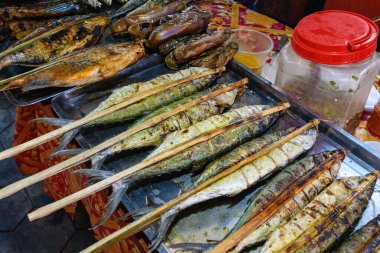 Kamboçya 'nın Kep kentindeki canlı bir markette metal tepsilerde sergilenen bambu çubuklarıyla ızgara balık seçimi. Görüntü kömürleşmiş dokuları, doğal renkleri ve tipik Güneydoğu sokak yemekleri ortamını vurguluyor