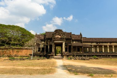Angkor Wat 'ın yıpranmış kumtaşı giriş köşkü açık güneş ışığında duruyor. Çerçevelenmiş kuru çimenler ve küçük bulutlarla dağılmış mavi gökyüzünün altındaki yapraklı ağaçlar. Süslü oymalar ve simetrik sütunlar tarihi Khmer anıtlarını vurguluyor