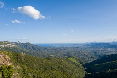 Geniş yeşil vadi uzak Akdeniz 'e doğru uzanır, engebeli dağ yamaçlarıyla çerçevelenmiş ve açık mavi bir gökyüzü altında dağınık bulutlar Baunei ve Dorgali, Sardinya yakınlarında. Güneş ışığı, değişken araziyi ve doğal dokuları vurgular.