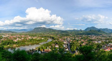 Luang Prabang şehri üzerinde Nam Khan nehri yemyeşil tepeleri ve kuzey Laos 'taki dramatik dağları sararken. Parlak gün ışığı ve dağınık bulutlar canlı tropikal manzarayı ve geleneksel çatıları vurgular..