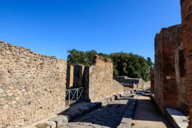 Pompeii arkeolojik sahasında yıpranmış taş ve tuğla duvarlarla çevrili tarihi bir Roma caddesinin manzarası. Sahnede sert duvarcılık, derin gölgeler ve parlak mavi gökyüzünün altında yemyeşil bir alan yer alıyor..