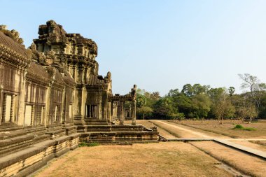 Angkor Vat, Kamboçya 'daki antik kumtaşı galerisinin yan görünüşü, sıcak güneş ışığı ile karışık Khmer oymaları ve balyalanmış pencereler. Tapınak yapısı kuru çimenli bir avluya ve tropik ağaçlarla çevrili bir toprak yola açılıyor.