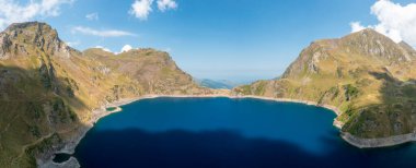 Pireneler 'deki Lac Bleu' nun geniş hava manzarası dik çimenli dağlar ve parlak mavi bir yaz gökyüzü altında dramatik kayalık yamaçlar tarafından çerçevelenmiştir. Derin mavi göl, güneşli alp desenleri ve dağınık bulutlarla çelişiyor..
