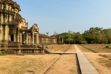 Angkor Vat, Kamboçya 'daki antik kumtaşı galerisinin yan görünüşü, sıcak güneş ışığı ile karışık Khmer oymaları ve balyalanmış pencereler. Tapınak yapısı kuru çimenli bir avluya ve tropik ağaçlarla çevrili bir toprak yola açılıyor.