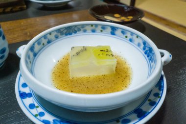 A small square of tofu topped with translucent kiwi jelly sits in a shallow pool of golden sauce, presented in a decorative blue and white bowl. The scene highlights smooth textures and a refined Japanese dining atmosphere.