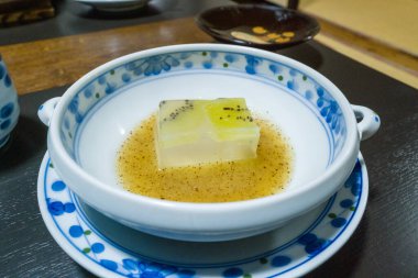 Üstü şeffaf kivi jölesiyle kaplı bir kare tofu, beyaz ve mavi desenli porselen bir kasede servis edilen kehribar sosunun sığ havuzunda oturur. Yakın plan parlak dokular ve rafine Japon yemek masası..