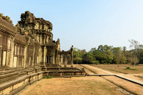 Angkor Vat, Kamboçya 'daki antik kumtaşı galerisinin yan görünüşü, sıcak güneş ışığı ile karışık Khmer oymaları ve balyalanmış pencereler. Tapınak yapısı kuru çimenli bir avluya ve tropik ağaçlarla çevrili bir toprak yola açılıyor.