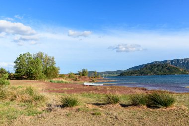Canlı kırmızı bir kıyı şeridi, dağınık otlar ve küçük beyaz bir teknenin yer aldığı Salagou Gölü 'nün geniş manzarası. Açık mavi gökyüzü ve uzak yeşil tepeler barışçıl bir göl atmosferi yaratır..