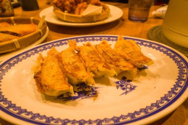 Altın tavada kızartılmış gyoza köfteleri, sıcak bir Osaka restoranında mavi ve beyaz seramik tabakta sıralanır. Sıcak ortam aydınlatması gevrek dokuyu ve davetkar sunumu geliştirir.