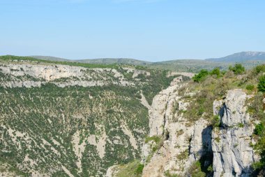 Navacelles Sirki 'ndeki engebeli bir vadiye tepeden bakan yeşil bitki örtülü dik kireçtaşı kayalıklar. Manzara güneşli, geniş ve doğal olarak berrak bir gökyüzünün altında..