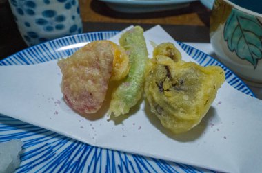 Japon sofra takımı ile çevrili mavi çizgili bir seramik tabağın üzerinde üç çeşit sebze parçası beyaz kağıda dizilmiştir. Yakın plan, rafine bir yemek ortamındaki altın, havadar ve ince renkleri vurguluyor..