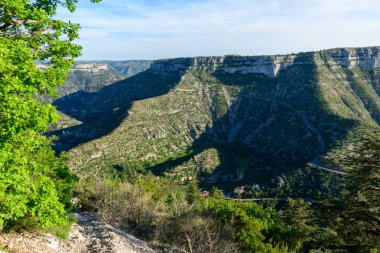 Engebeli kireçtaşı kayalıkları ve Cirque de Navacelles 'deki derin, gölgeli bir vadi, canlı yeşil yapraklar ve parlak mavi bir gökyüzü altında. Manzarada çarpıcı jeolojik oluşumlar ve bereketli Akdeniz bitkileri yer alıyor..