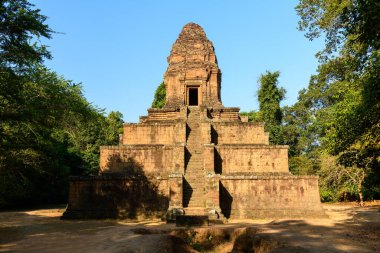 Phnom Bakheng 'in yıpranmış kumtaşı piramidi Angkor, Kamboçya' daki tropik ormanın üzerinde yükselir. Kıtır güneş ışığı, açık mavi gökyüzünün altındaki basamaklı mimari ve dokulu taş işçiliğini gözler önüne seriyor..