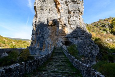 Ancient stone pathway ascends toward a towering limestone cliff with a natural cave opening, surrounded by autumn foliage in the Vikos Gorge region of Epirus, Greece. Bright sunlight highlights the textured rock face and rugged landscape under a