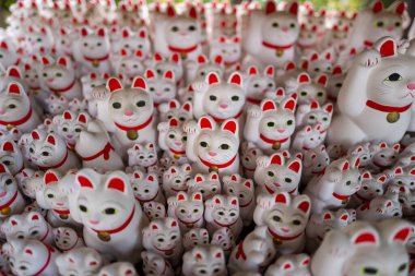 Tokyo 'daki Gotokuji Tapınağı' nda canlı bir şekilde sergilenen, kırmızı detayları ve altın çanları olan beyaz şanslı kedi heykellerinin yoğun şekilde gruplandırılması. Görüntü, yukarıdan tekrarlama, oyuncu ifadeler ve yumuşak gün ışığı süzgeçlerini vurgular.