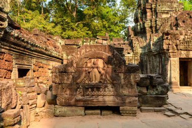 Mitolojik figürlerin yer aldığı entrika ile oyulmuş kumtaşı lintel, Kamboçya Angkor bölgesindeki Ta Som tapınağının yıpranmış kalıntıları arasında duruyor. Sıcak öğleden sonra güneşi, bereketli tropikal kuşaklar tarafından çerçevelenmiş, ayrıntılı kaya döşemeleri ve dokulu taş işçiliğini ortaya çıkarır.