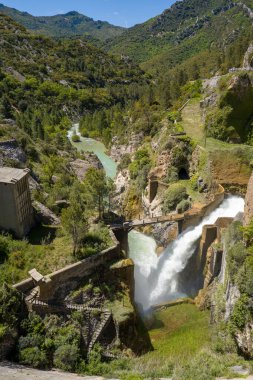 Barajdan derin bir vadiye güçlü bir su akıntısı yükseliyor. Etrafı yemyeşil tepeler ve Las Penas 'taki engebeli dağlarla çevrili. Güneş ışığı taşın, betonun ve canlı bahar bitkisinin dokusunu vurgular.