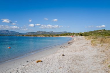 Spiaggia la Salinedda, Sardinya 'daki geniş kumlu kıyı şeridi şeffaf mavi deniz, uzak dağ sıraları ve canlı bir gökyüzü altında dağınık bulutlar..