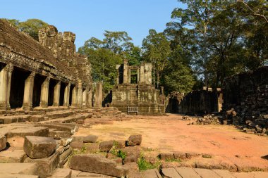 Altın öğleden sonra ışığı, Angkor, Kamboçya 'daki Bayon Tapınağı' nın açık avlusunda ve yıpranmış taş sütununda yanar. Eski kumtaşı blokları ve kısmen harap olmuş duvarlar yüksek orman ağaçlarının arasında duruyor, sakin bir tarihi sahne yaratıyor..