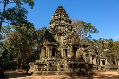 The central sandstone sanctuary of Thommanon temple stands illuminated by afternoon light, surrounded by tall trees and deep shadows in Angkor, Cambodia. Weathered carvings and layered stonework are highlighted beneath a clear blue sky, set against