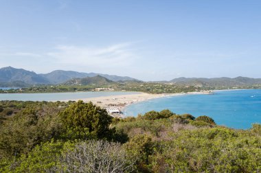 Turkuaz bir deniz ile Porto Giunco 'daki sakin bir göl arasındaki geniş kumlu kıyı şeridi kıvrımları. Berrak bir gökyüzü altında yemyeşil tepeler ve uzak dağlarla destekleniyor..