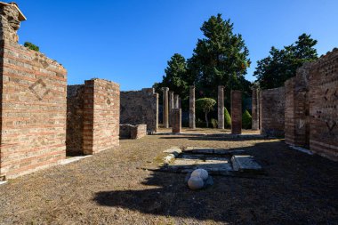 Antik tuğla ve volkanik taş duvarlar İtalya 'nın Pompeii kentindeki arkeolojik kamaralarda güneşli bir sütun ve açık bir avlu oluşturuyor. Geometrik duvarcılık desenleri ve yıpranmış sütunlar, Akdeniz yeşilliğinin ötesinde, canlı mavi bir gökyüzünün altında duruyor..
