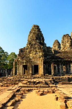 Angkor, Kamboçya 'daki Bayon Tapınağı' nın girişinde heybetli yüz oymalarına sahip antik taş kule yükseliyor. Sıcak güneş ışığı, yıpranmış kumtaşını ve karmaşık Khmer detaylarını vurguluyor. Orman ağaçları ve tarihi gökyüzünü çevreleyen parlak mavi bir gökyüzü.