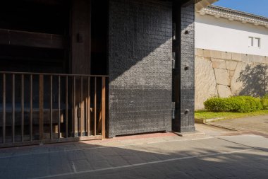 Osaka şatosunun girişinde demir kapılar kısmen açık, güneş ışığı taş kaldırımlara ve tarihi duvarlara keskin gölgeler bırakıyor. Sahnede açık mavi gökyüzü altında desenli metal, ahşap ve taş elementler yer alıyor..