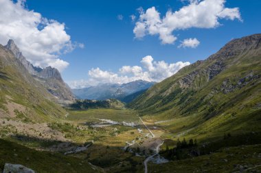 Col de la Seigne yakınlarındaki İtalyan Alpleri 'nde geniş yeşil bir vadi, engebeli dağlarla çevrili ve dağınık mavi bulutlarla dolu parlak mavi bir gökyüzünün altında dolambaçlı bir nehir yatağı.