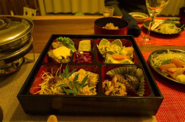 Elegant assortment of Japanese dishes arranged in a lacquered bento box, surrounded by side plates and glassware on a warmly lit dining table. The meal features a variety of textures, colors, and fresh ingredients for a refined culinary presentation.