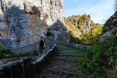 Yunanistan 'ın Epirus kentindeki Pont Gefiri Kokkorou kenti yakınlarında tarihi taş kemer köprüsü yüksek kireçtaşı uçurumlarının altındaki kayalık bir nehir yatağını ve sonbahar yapraklarını geçer. Yumuşak gün ışığı, desenli duvarcılık, canlı yeşillik ve dramatik kanyon manzarasını vurgular.