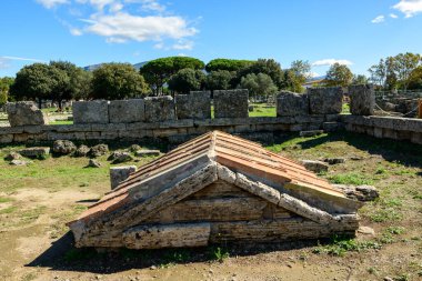 Üçgen çatılı yıpranmış taş bir mezar İtalya 'nın Paestum kentindeki antik taş bloklar arasında yer alıyor. Sahne, yemyeşil ağaçlar ve arka planda canlı mavi bir Akdeniz gökyüzü ile canlı güneş ışığı altında çekilmiştir..