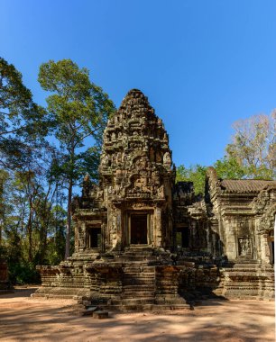 The main sandstone tower of Thommanon temple rises under a clear blue sky, surrounded by tall forest trees in Angkor, Cambodia. Crisp sunlight highlights the intricate carvings and weathered stonework, casting deep shadows on the sandy ground.