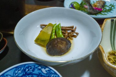 A white ceramic bowl holds simmered shiitake mushroom, green beans, bamboo shoot, and wild mountain vegetables in a light broth. The arrangement features earthy colors and delicate textures under gentle lighting.