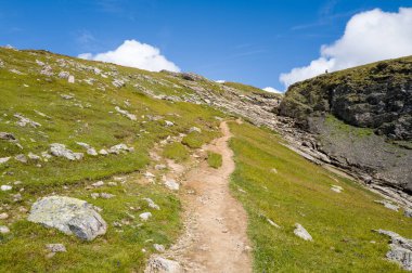 Çimenli bir dağ yamacında dar bir toprak yol kıvrılır taşlarla ve engebeli kayalarla dağılır parlak mavi gökyüzünün altında kabarık bulutlarla.