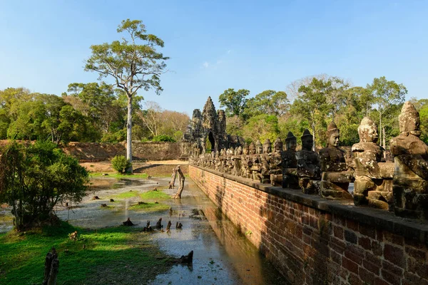 Kamboçya 'daki Siem Reap yakınlarındaki Angkor Thom' un anıtsal giriş kapısına uzanan köprüye uzanan uzun sıra antik taş koruyucu figürler. Tarihi Khmer heykelleri, bereketli tropikal ağaçları ve sığ hendekleri olan ılık gün ışığıyla aydınlatılır.