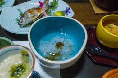 İnce beyaz balık sashimi dilimleri, parlak mavi bir seramik kasede deniz yosunu garnitürü ile servis edilir, koyu bir yemek masasında çeşitli renkli Japon yemekleri ile çevrilidir. Düzenleme, taze dokuları ve zarif, davetkar...