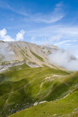 Canlı yeşil çayırlar, Beaufortain Alplerinde engebeli bir kireçtaşı sırtına doğru sürükleniyor, bulutların zerreleri yukarıdaki parlak yaz gökyüzünde sürükleniyor..
