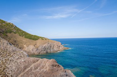 A rugged, rocky slope with patches of green vegetation meets the vivid blue Mediterranean Sea near Argentiera, Sardinia. Bright sunlight and a cloudless sky emphasize the wild coastal landscape and transparent water.