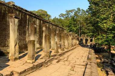 Bir sıra yıpranmış taş sütun, Angkor, Kamboçya 'daki Bayon Tapınağı' nda ustalıkla oyulmuş bir levrek yardım duvarının yanında güneşli bir yürüyüş yolu çiziyor. Tarihi Khmer kalıntıları, berrak mavi gökyüzünün altında gür ormanlarla çevrilidir.