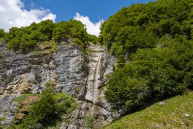 İnce bir şelale engebeli bir kireçtaşı uçurumu boyunca iniyor, yoğun yeşil ağaçlarla çerçeveleniyor ve Cirque du Fer-a-Cheval 'de gün ışığıyla aydınlatılıyor..