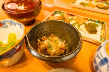 A serving of chilled tofu garnished with bonito flakes and scallions sits in a dark ceramic bowl, surrounded by assorted Japanese breakfast dishes on a wooden table. Warm lighting enhances the inviting textures and colors of the traditional setting.
