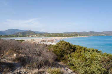 Expansive sandy shoreline meets a turquoise bay and tranquil lagoon at Porto Giunco, with green hills and distant mountains under a clear blue sky.